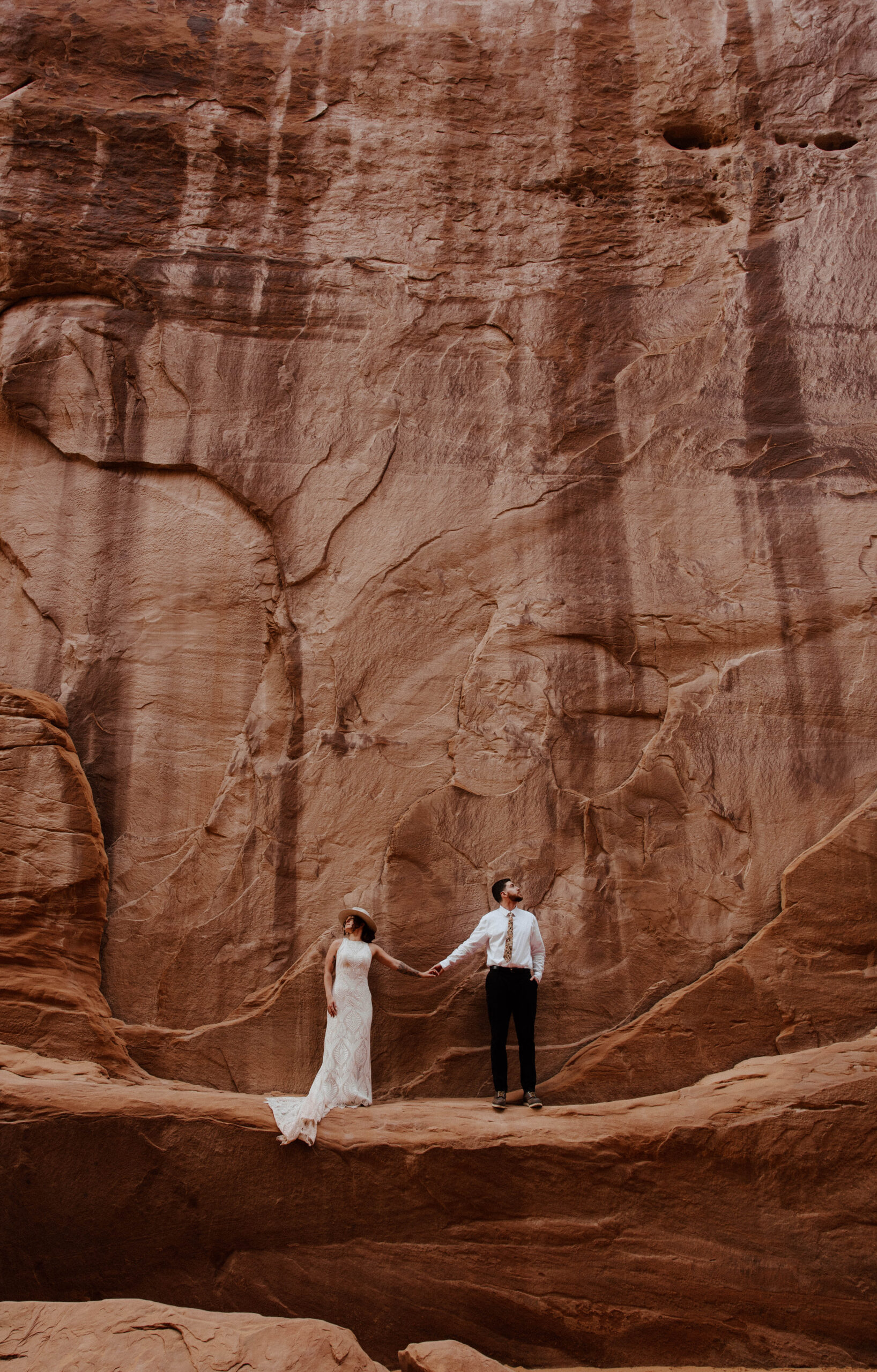 Couple standing in shaded rock alcove on cliff surrounded by red rock formations during Moab elopement.